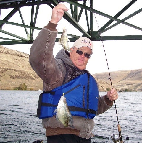 An angler showcasing two crappies caught on jigs below a bridge.